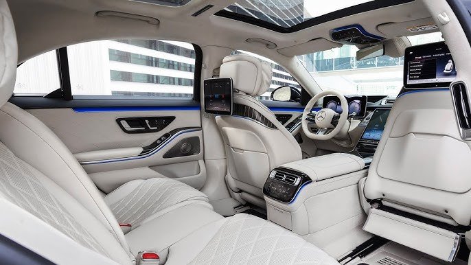Luxurious interior of a white Mercedes-Benz S-Class featuring premium quilted leather seats, dual entertainment screens, and blue ambient lighting.