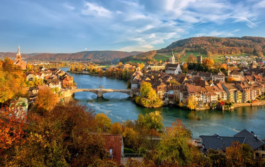 Aerial panoramic view of a picturesque Swiss town on the banks of the Rhine River during autumn, with colorful trees and a historic stone bridge.